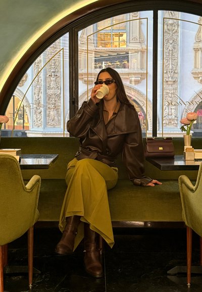 Mujer con gafas de sol, chaqueta de cuero y falda larga sentada en un banco verde en una cafetería, sosteniendo una taza, con una ventana ornamentada detrás.