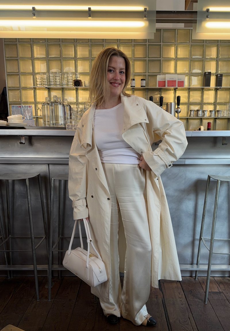 Femme en manteau et pantalon blancs tenant un sac à main blanc, debout et souriante dans un café moderne avec des tabourets en métal et des étagères murales en verre derrière elle.