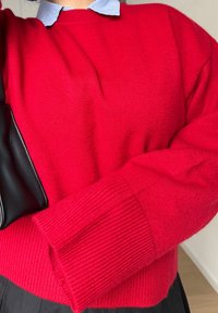 Crveni pleteni džemper s rebrastim manšetama i okruglim izrezom, slojevit preko svijetloplave košulje s ovratnikom. Crna torba vidljiva je sa strane.