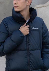 Casaco acolchoado preto da Columbia com colarinho alto, fecho de correr e costuras nos ombros. Apresenta textura volumosa e logótipo no lado esquerdo.