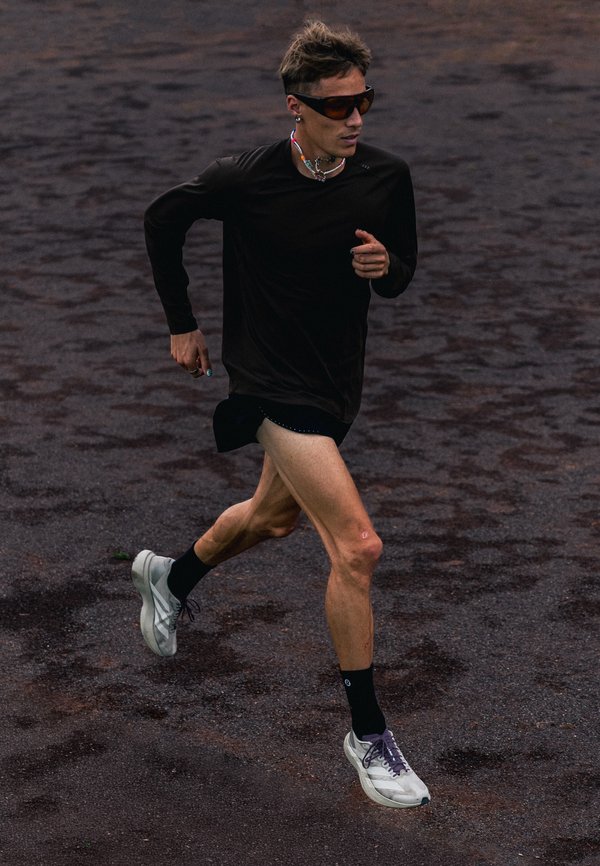 Coureur portant un t-shirt noir à manches longues, un short, des lunettes de soleil et des baskets blanches, jogging sur une surface texturée foncée en plein air.