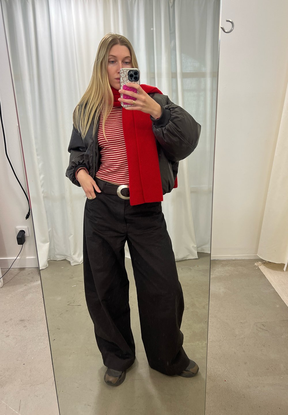 Calças largas pretas, cachecol vermelho com textura em costelas, camisola listrada em vermelho e branco, e uma jaqueta preta acolchoada. Fivela de cinto refletiva. Cenário de espelho.
