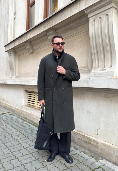 Hombre con abrigo oscuro y gafas de sol sostiene una bolsa negra, de pie sobre una calle adoquinada frente a un edificio de piedra beige con elementos decorativos.