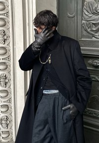 Man in zwarte jas, handschoenen en grijze broek staat bij een sierlijke muur, bedekt een deel van zijn gezicht met één hand en kijkt naar beneden.