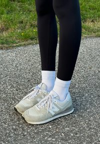 Baskets New Balance gris clair avec des lacets et des accents blancs, portées avec des chaussettes blanches mi-mollet et des leggings noirs sur une surface texturée.