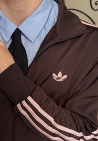 Personne portant une chemise bleu à col et une cravate noire sous une veste Adidas marron avec des rayures et un logo roses, bras croisés sur la poitrine.