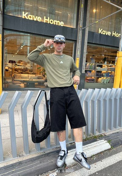 Pantalones cortos negros, camisa de manga larga oliva con logo, gafas de sol, zapatillas plateadas, calcetines blancos y bolso negro de hombro. Entorno urbano.