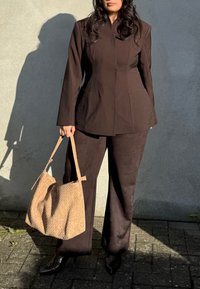 Brun skræddersyet blazer med matchende skræddersyede bukser, parret med en vævet beige håndtaske og spidse sorte hæle, sat mod en neutral baggrund.