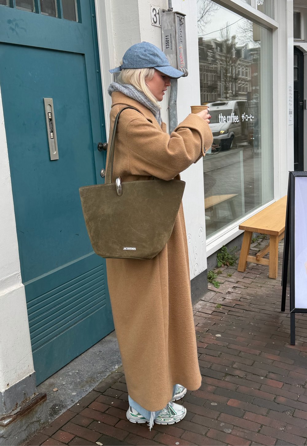 Femme portant un long manteau camel et une casquette bleue, tenant une tasse de café, debout devant un café avec un grand sac fourre-tout vert.