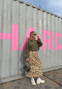 Zeleni flis pulover, khaki kamuflažni suknja, bijele tenisice i mala zelena torba. Roze grafit na sivom kontejneru u pozadini.