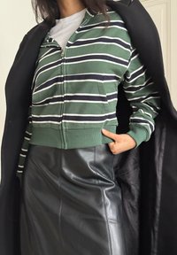 Personne portant un sweat à capuche zippé à rayures vertes, blanches et noires par-dessus un t-shirt blanc, une jupe en cuir noire, et un manteau noir posé sur les épaules.