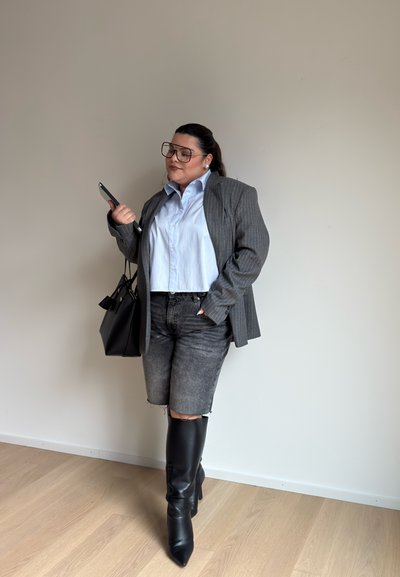 Blazer gris a rayas finas sobre una camisa de color azul claro con cuello, acompañado de shorts de mezclilla negra desgastados y botas de tacón alto negras hasta la rodilla.