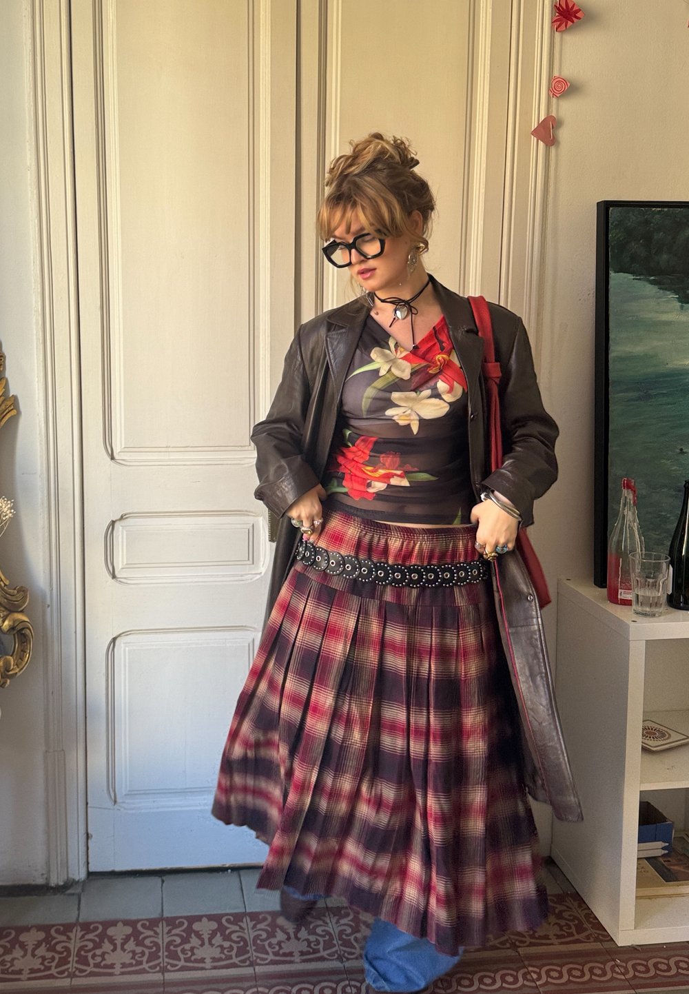 Καφέ δερμάτινο παλτό πάνω από ένα μαύρο φλοράλ τοπ, φούστα πλισέ σε κόκκινο και μαύρο καρό, διακοσμημένη ζώνη και μπλε τζιν ορατό στο κάτω μέρος.