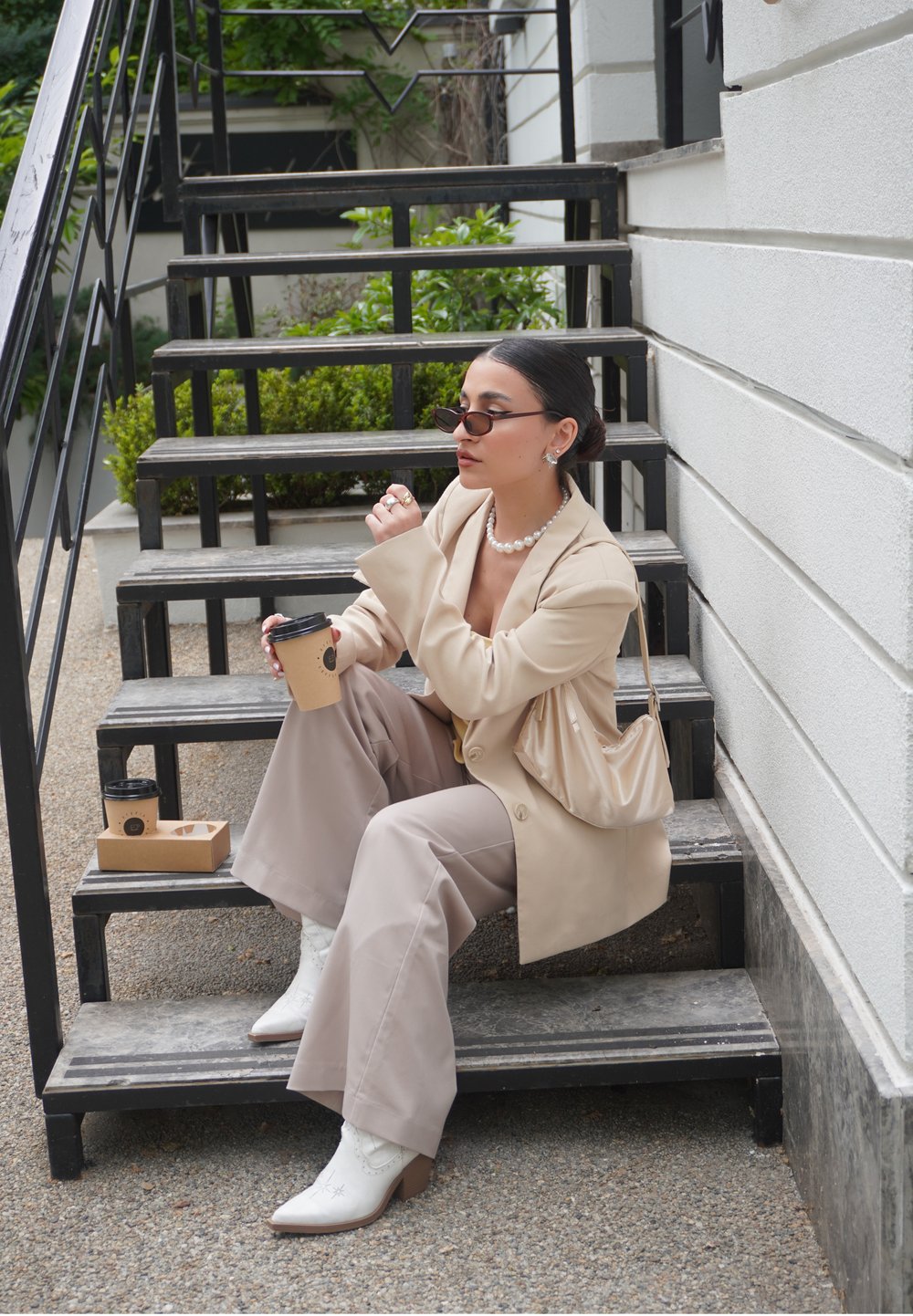 Bež blazer na bijeloj majici, široke bež hlače, bijele čizme u western stilu. Crne sunčane naočale, biserna ogrlica, drži šalicu kave. Drvene stepenice.