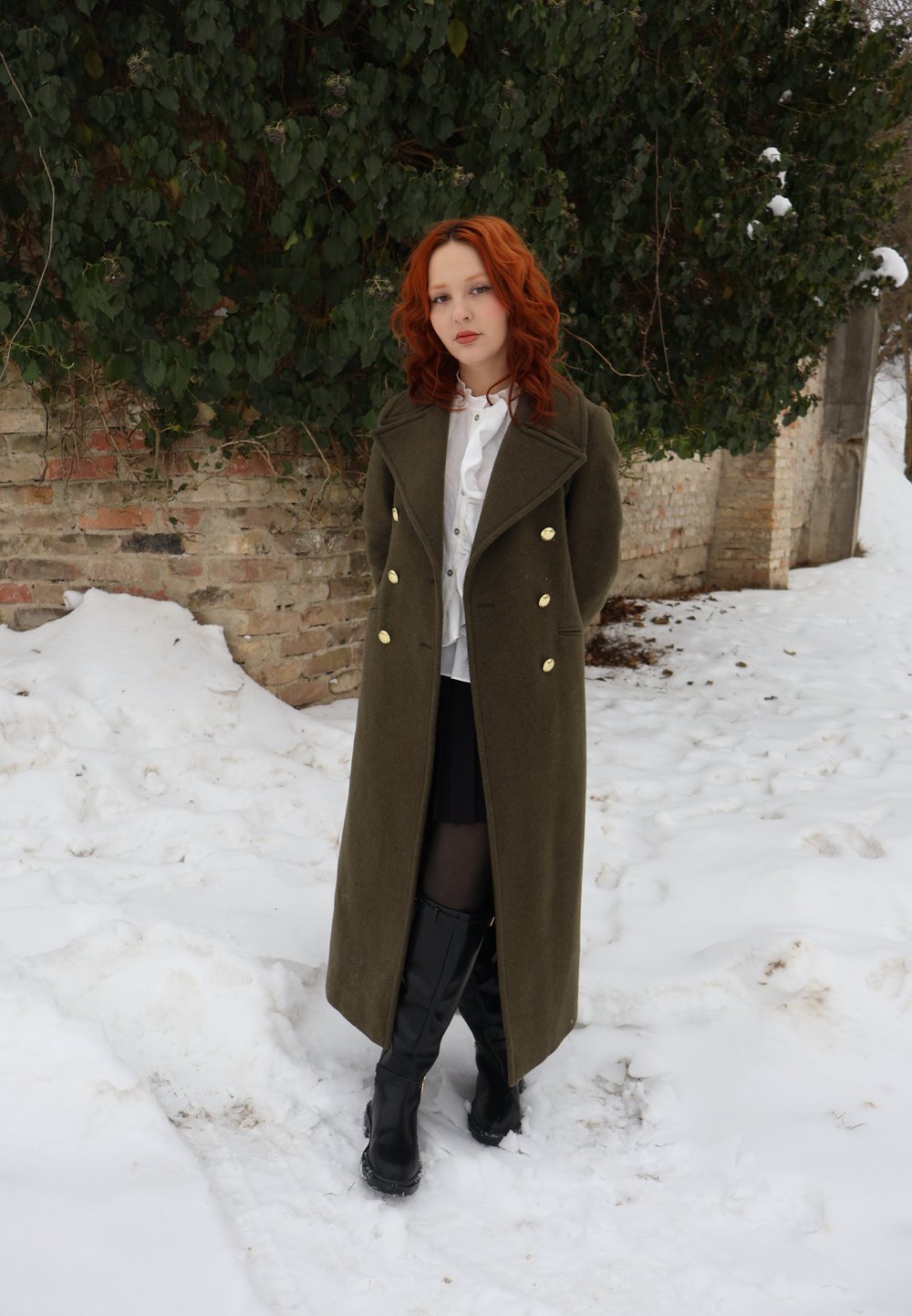 Jeune femme aux cheveux roux portant un long manteau olive, des bottes noires et un chemisier blanc, debout sur la neige devant un mur de briques recouvert de lierre.