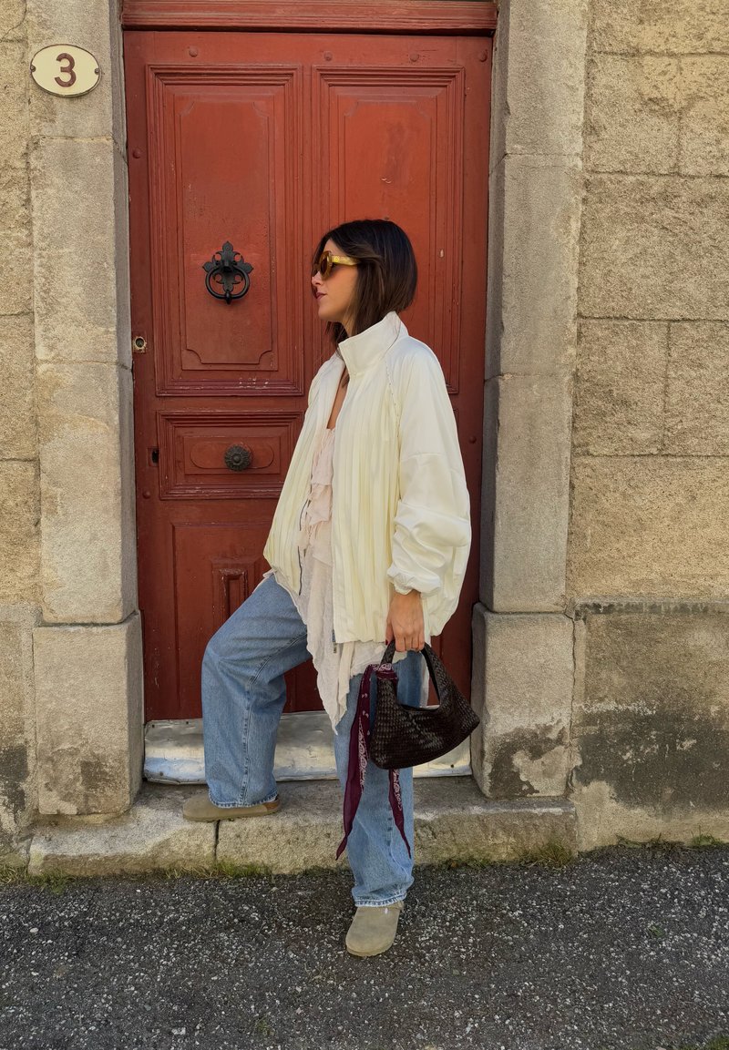 Bomberjacke in hellem Creme, darüber eine weiße Bluse, verwaschene blaue Jeans, graue Clogs und eine kleine dunkle geflochtene Handtasche mit einem gemusterten Schal. Rote Tür im Hintergrund.