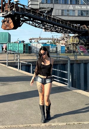 Top negro sin mangas, pantalones cortos de denim desgastados y botas negras. Cabello largo y negro, múltiples tatuajes en las piernas. Entorno industrial con maquinarias de fondo.