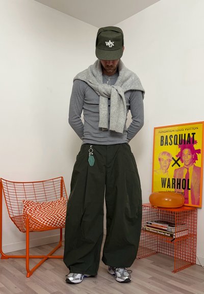 Hombre con pantalones oscuros extragrandes, camisa de manga larga gris, suéter gris atado alrededor de los hombros, gorra verde, de pie entre muebles naranjas y un cartel colorido.