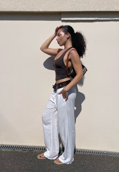 Top de tirantes marrón ajustado y pantalones de lino blancos sueltos, complementados por un bolso oscuro con una correa naranja. De pie contra una pared clara.