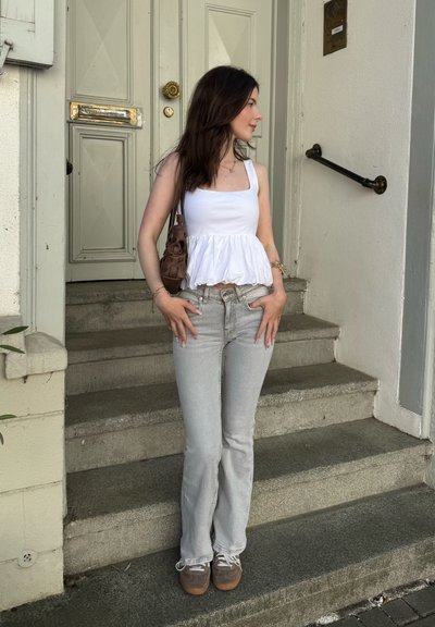 Top peplum blanco, jeans acampanados gris claro, bolso marrón y zapatillas gris; de pie en unos escalones de piedra frente a una puerta verde.