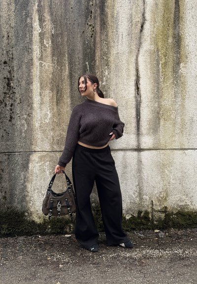 Mujer de pie junto a una pared texturizada, vistiendo un suéter oscuro sin hombros, pantalones anchos negros, sosteniendo un bolso marrón y mirando hacia su izquierda.