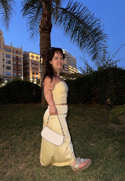 Mujer con top sin tirantes y falda amarillo pálido sosteniendo bolso blanco, de pie sobre la hierba junto a una palmera con edificios iluminados al anochecer.