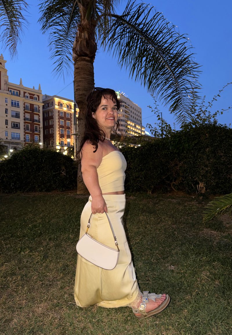 Femme en haut bustier jaune pâle et jupe tenant un sac blanc, debout sur l'herbe près d'un palmier avec des bâtiments illuminés au crépuscule.