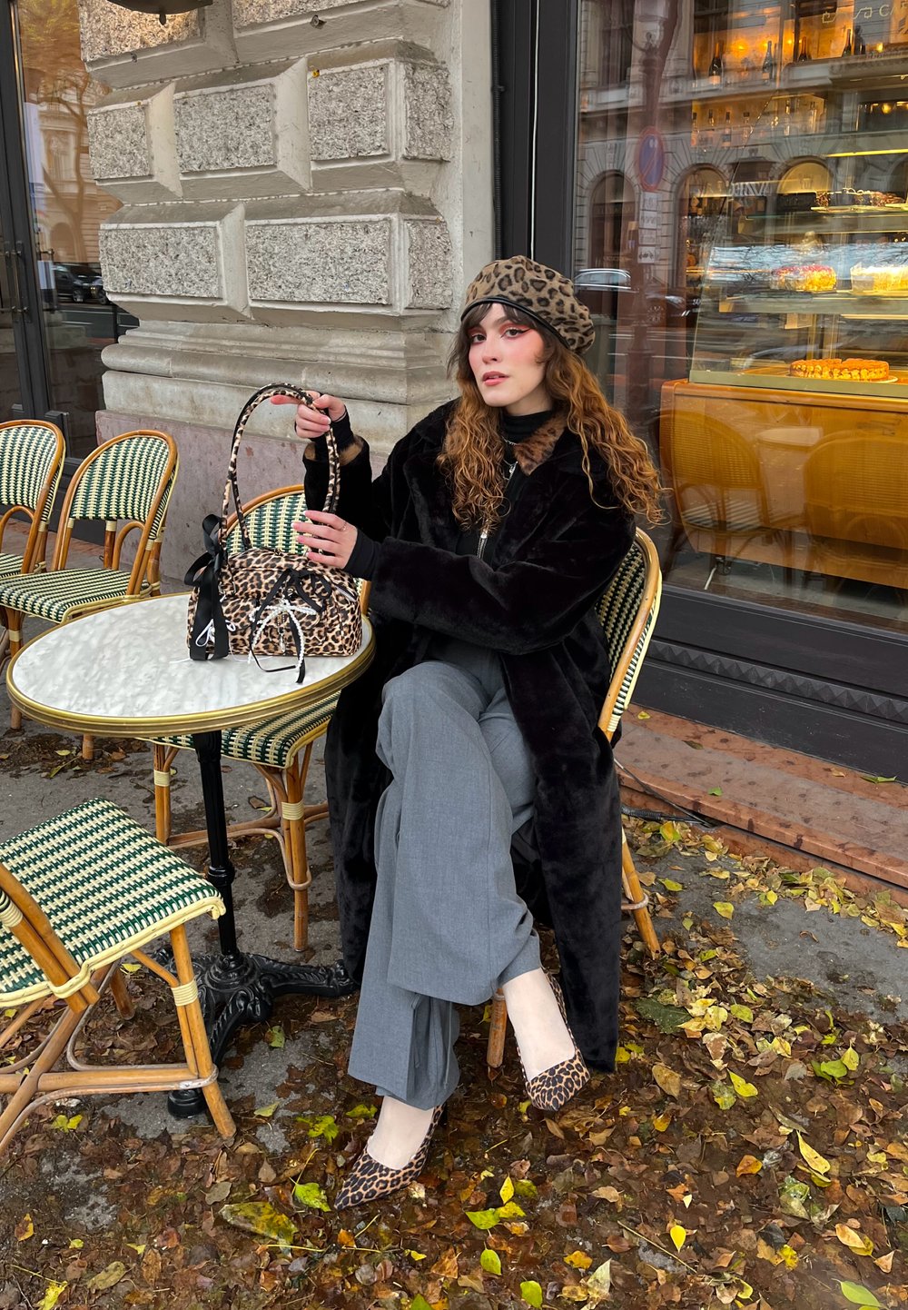 Borsa a stampa leopardata, cappotto in pelliccia sintetica nera, pantaloni grigi a gamba larga, scarpe basse a stampa animale, seduta a un tavolo di bistrò con sedie verdi intrecciate.