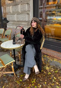 Borsa a stampa leopardata, cappotto in pelliccia sintetica nera, pantaloni grigi a gamba larga, scarpe basse a stampa animale, seduta a un tavolo di bistrò con sedie verdi intrecciate.