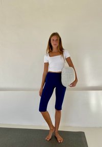 Witte fitted crop top met korte mouwen, gecombineerd met marineblauwe capri leggings. Houdt een grijs en wit geruite tas vast. Effen achtergrond.