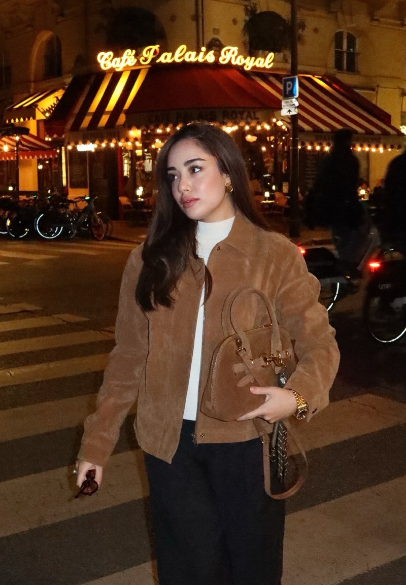 Brown suede jacket over a white turtleneck, black wide-leg pants, holding a small brown handbag with gold hardware, nighttime street setting.