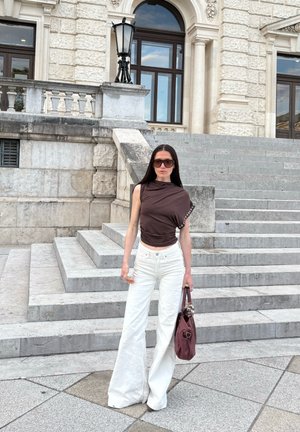 Mujer con cabello largo y oscuro, que lleva una camiseta sin mangas marrón oscuro, pantalones blancos de pierna ancha, gafas de sol, sosteniendo un bolso granate, de pie sobre escalones de piedra.