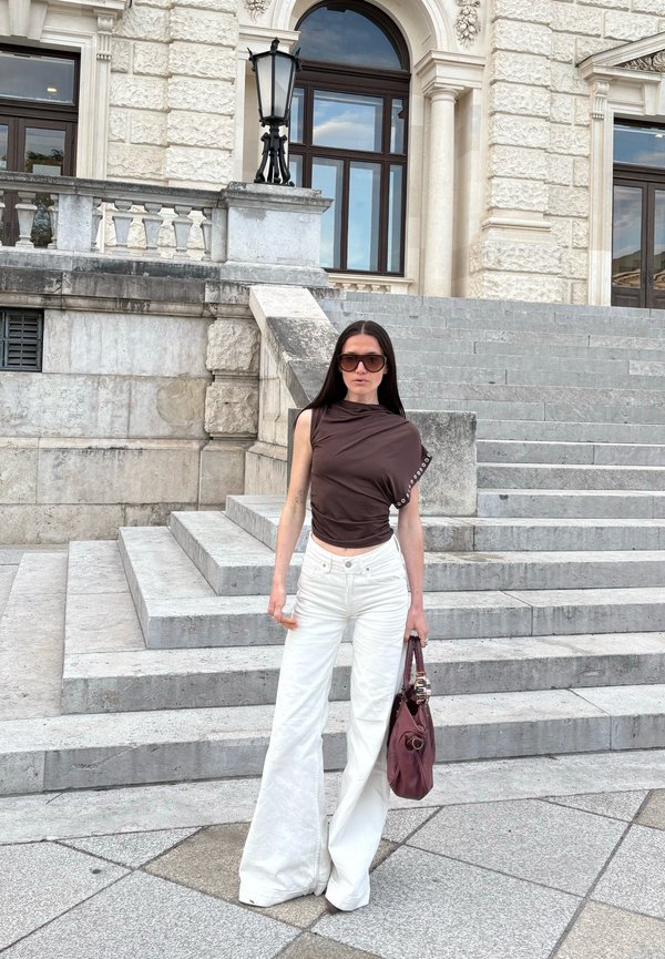 Femme aux longs cheveux foncés portant un haut sans manches marron foncé, un pantalon blanc à jambes larges, des lunettes de soleil, tenant un sac à main bordeaux, debout sur des marches en pierre.