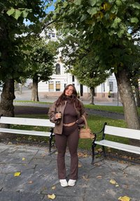 Brązowa skórzana kurtka na ciemnym brązowym dopasowanym topie i spodniach dzwonach. Białe trampki, trzyma filiżankę kawy, z brązową torebką. W tle zielone drzewa.