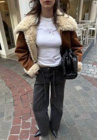 Femme debout sur une rue pavée, portant une veste en peau lainée marron, un haut blanc ajusté, un jean noir ample, des chaussures noires pointues, tenant une pochette noire texturée.