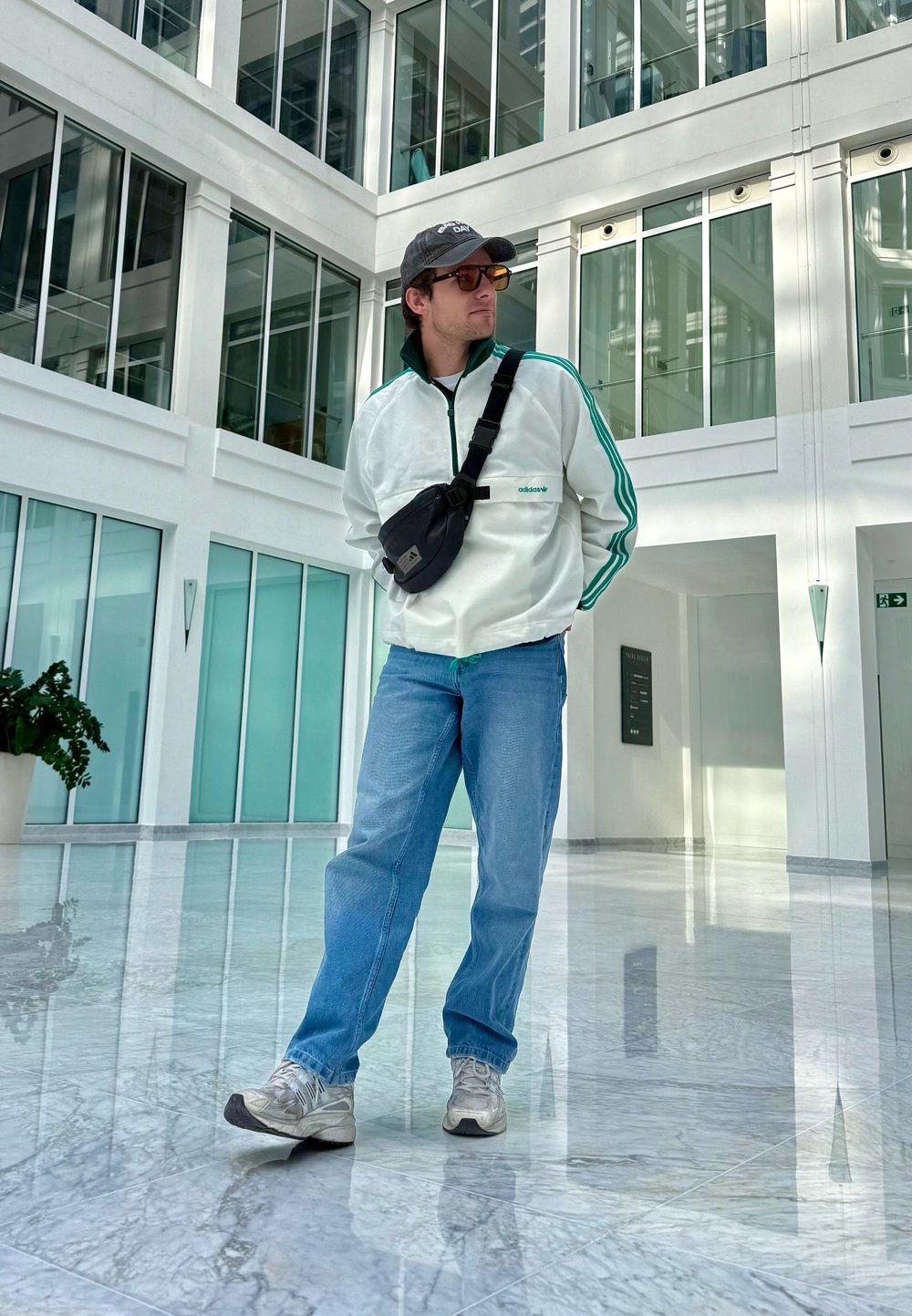 Casaco branco com fecho de correr e riscas verdes, jeans azuis e ténis prateados; mala cruzada preta e boné; interior moderno com paredes de vidro.