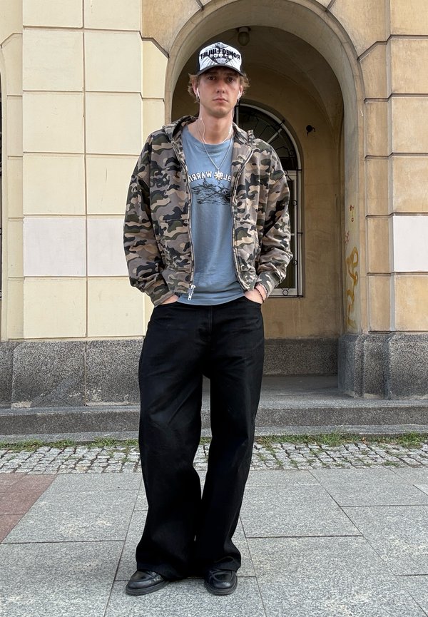 Un jeune homme se tient debout, les mains dans les poches, portant une veste camouflage, un t-shirt bleu, un pantalon noir à jambes larges, des chaussures noires et une casquette blanche en extérieur.