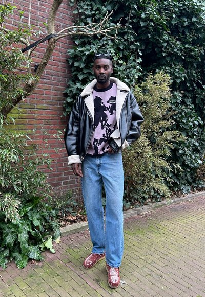 Chaqueta de cuero sintético negra con cuello de borrego, sudadera morada con estampado, jeans azules claros y zapatos rojos con estampado. Fondo de follaje verde.