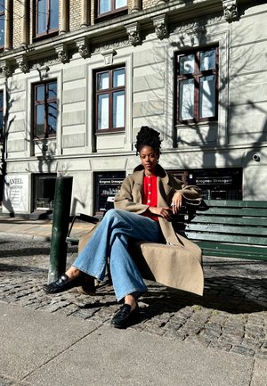 Modelo sentado en un banco verde, vistiendo un abrigo marrón, una camiseta roja y pantalones vaqueros de pierna ancha azules. Zapatos negros. Fondo urbano con pavimento de piedra.