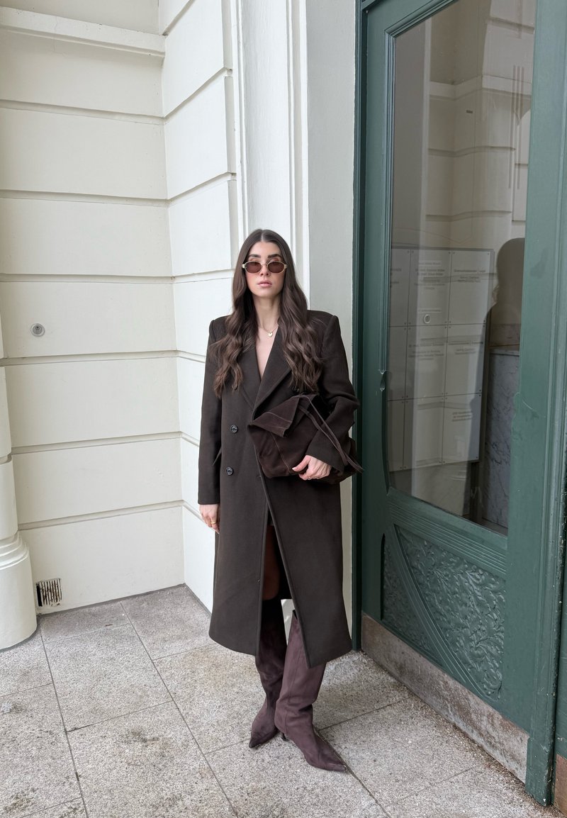 Femme aux longs cheveux noirs portant des lunettes de soleil, un long manteau marron foncé, des bottes assorties, tenant un sac marron foncé, debout près d'une porte verte.