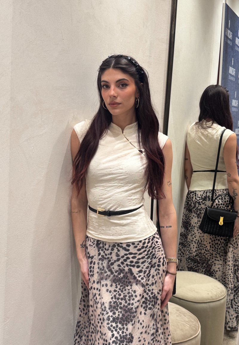 Kobieta o długich ciemnych włosach stoi przy białej ścianie, ubrana w białą bluzkę bez rękawów, spódnicę w panterkę, czarny pasek i trzyma czarną torebkę.