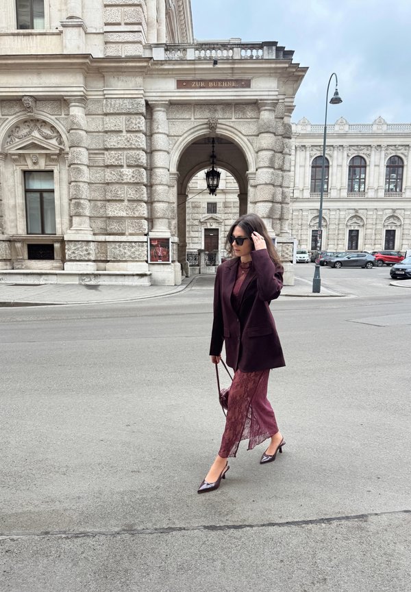 Ženska z zaščitnimi sončnimi očali, oblečena v temno vijoličast blazer in čipkasto obleko, hodi po mestni ulici ob okrašeni kamniti stavbi pod oblačnim nebom.