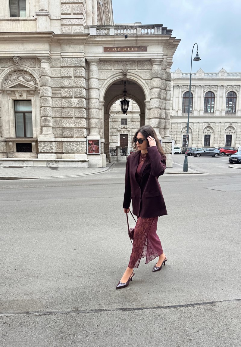 Γυναίκα με γυαλιά ηλίου, φοράει σκούρο μωβ σακάκι και δαντελένιο φόρεμα, περπατά σε δρόμο της πόλης κοντά σε διακοσμημένο πέτρινο κτίριο κάτω από συννεφιασμένο ουρανό.