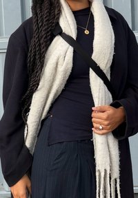 Crna majica s dugim rukavima nošena ispod debele, kremaste marame; uparena s hlačama na pruge. Marama ima obrubljene rubove. Zlatna ogrlica s okruglom privjeskom.