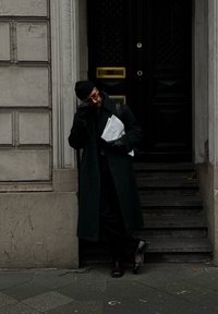 Μαύρο παλτό, μαύρο παντελόνι και καφέ παπούτσια. Κρατώντας εφημερίδες; φοράει μαύρα γάντια και σκουφί, στέκεται δίπλα σε μια σκοτεινή πόρτα.