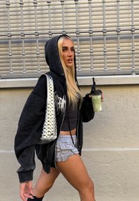Mujer rubia con sudadera negra, top corto y pantalones cortos grises con volantes, caminando junto a una ventana con barrotes de metal, sosteniendo una bebida verde con una pajita negra.