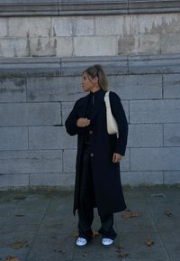 Abrigo largo negro con grandes botones, llevado sobre una blusa negra y pantalones de cuero. Bolso beige con superficie texturizada, zapatos blancos con patrones.