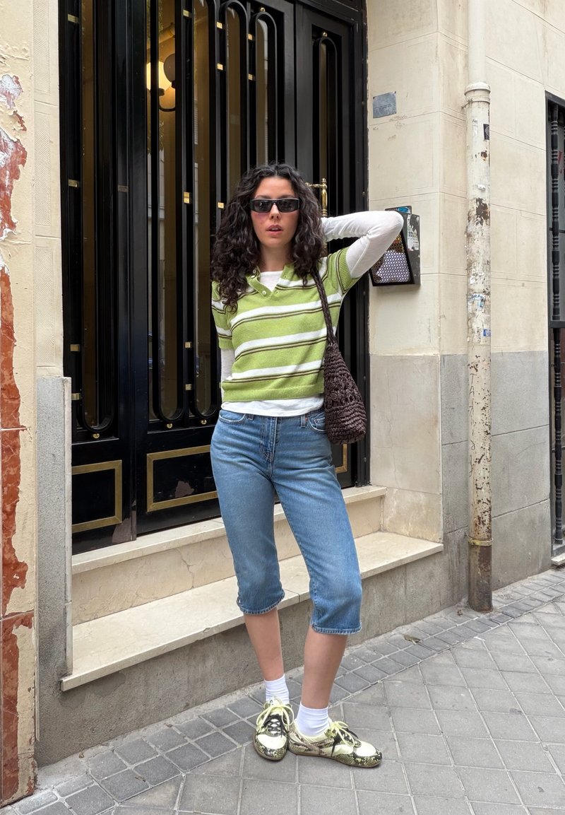 Jeune femme aux cheveux bouclés, lunettes de soleil, chemise verte rayée, pantacourt en jean et baskets, debout sur le trottoir de la ville devant une porte noire et dorée.
