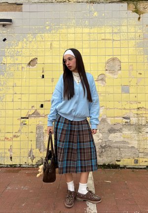 Joven con cabello largo, gafas, cárdigan azul, falda de cuadros y zapatos con estampado de leopardo, de pie frente a una pared amarilla con azulejos desgastados.