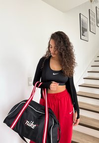 Femme en brassière de sport noire, veste noire et pantalon rouge Nike tenant un sac de sport Nike noir et rouge, debout près d'un escalier.